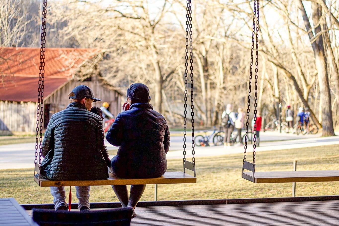 Two people in winter jackets sitting on wooden swings at Airship Coffee overlooking Coler Mountain Bike Preserve with bare trees and buildings in the background during fall