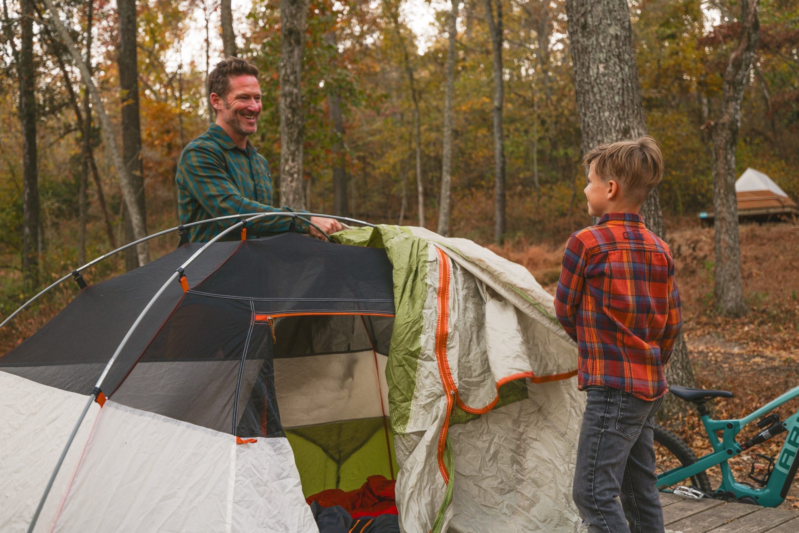 Smiling father in plaid shirt and young boy in flannel setting up gray and green camping tent among fall trees