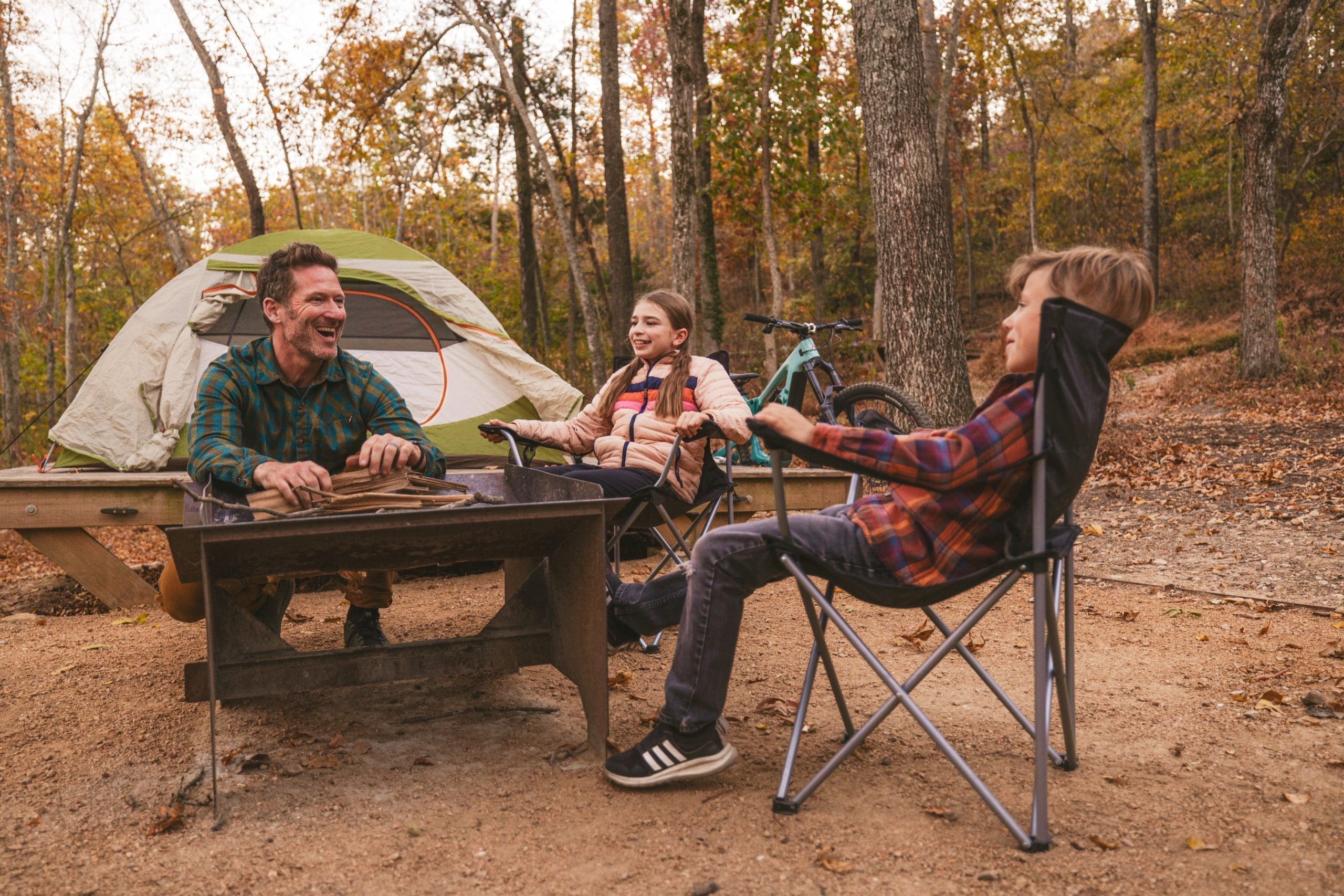 Father and two children laughing together at campsite picnic table with tent and mountain bikes visible in background among autumn trees