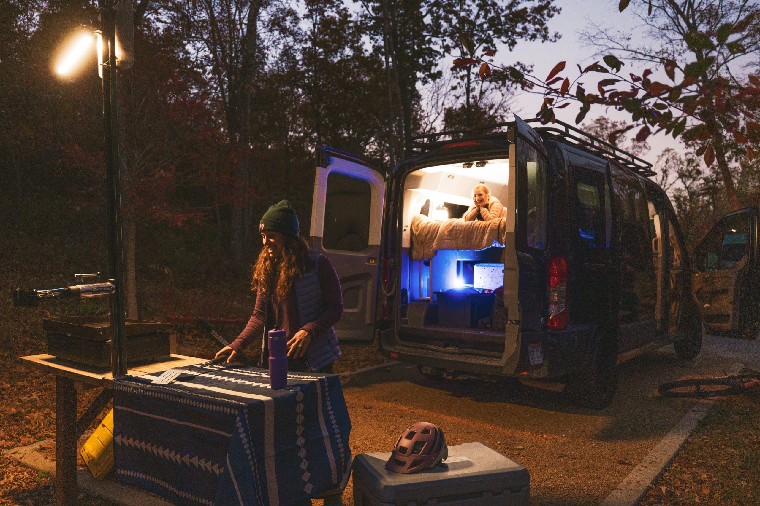 Woman in beanie organizing camping gear next to illuminated RV at dusk, with another person visible inside the lit RV interior