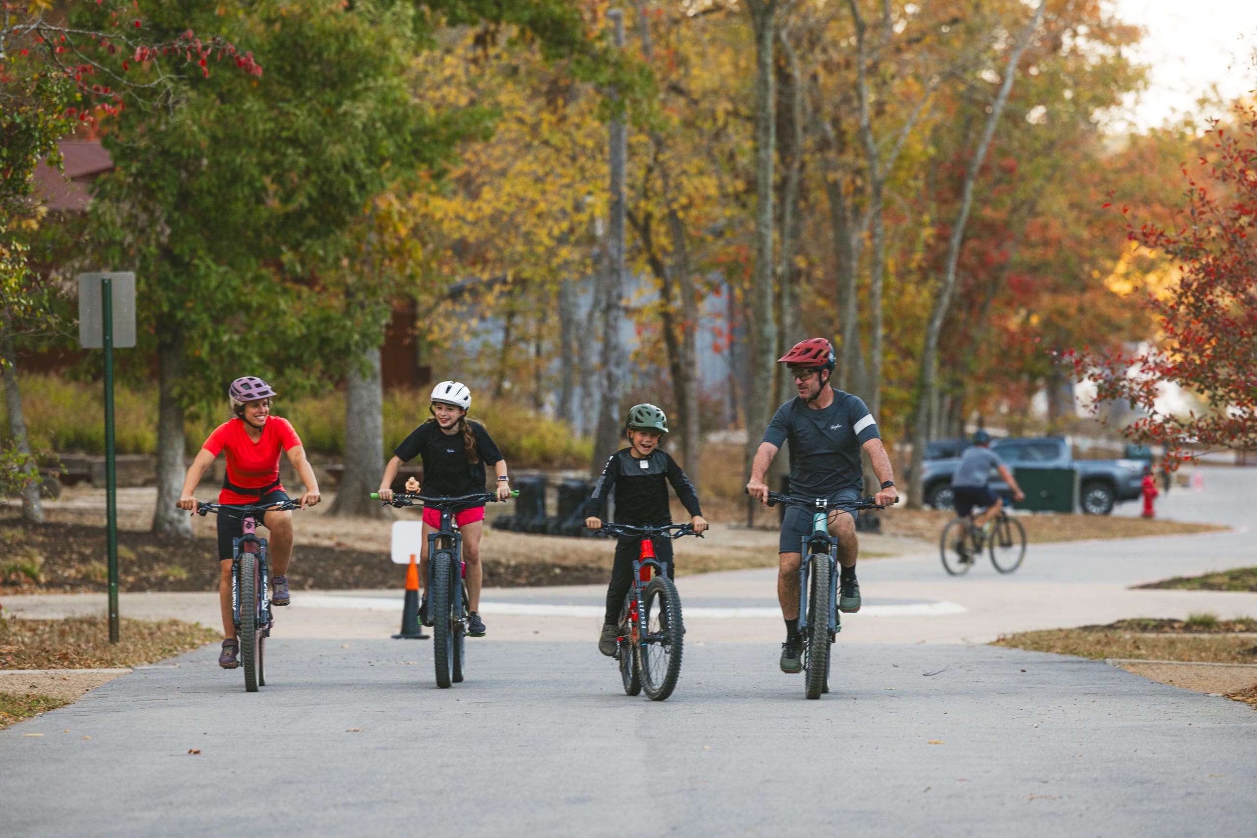 Family of four wearing helmets riding mountain bikes on paved trail through fall foliage, with vibrant autumn trees in background